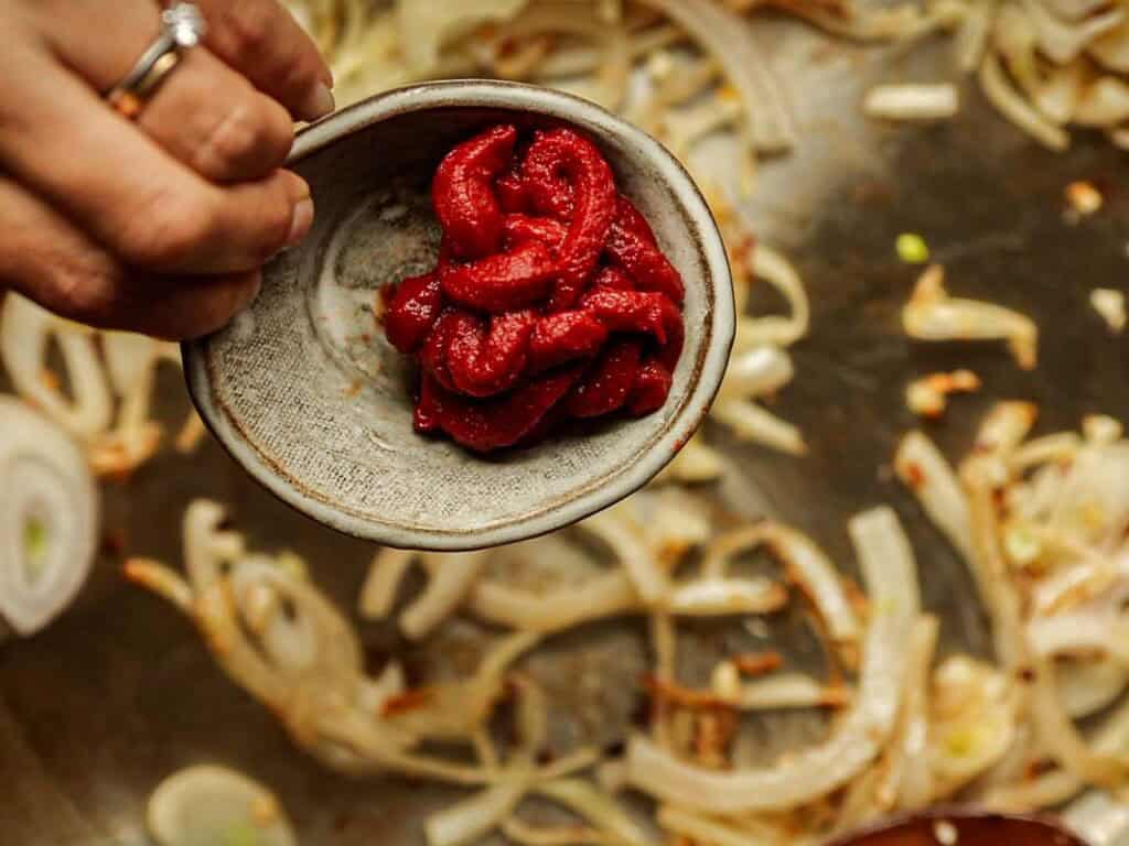 A hand holding a small ceramic bowl filled with red tomato paste, positioned above a pan with sautéing onions. The onions are browning and slightly translucent. The hand is adorned with rings on the fingers.